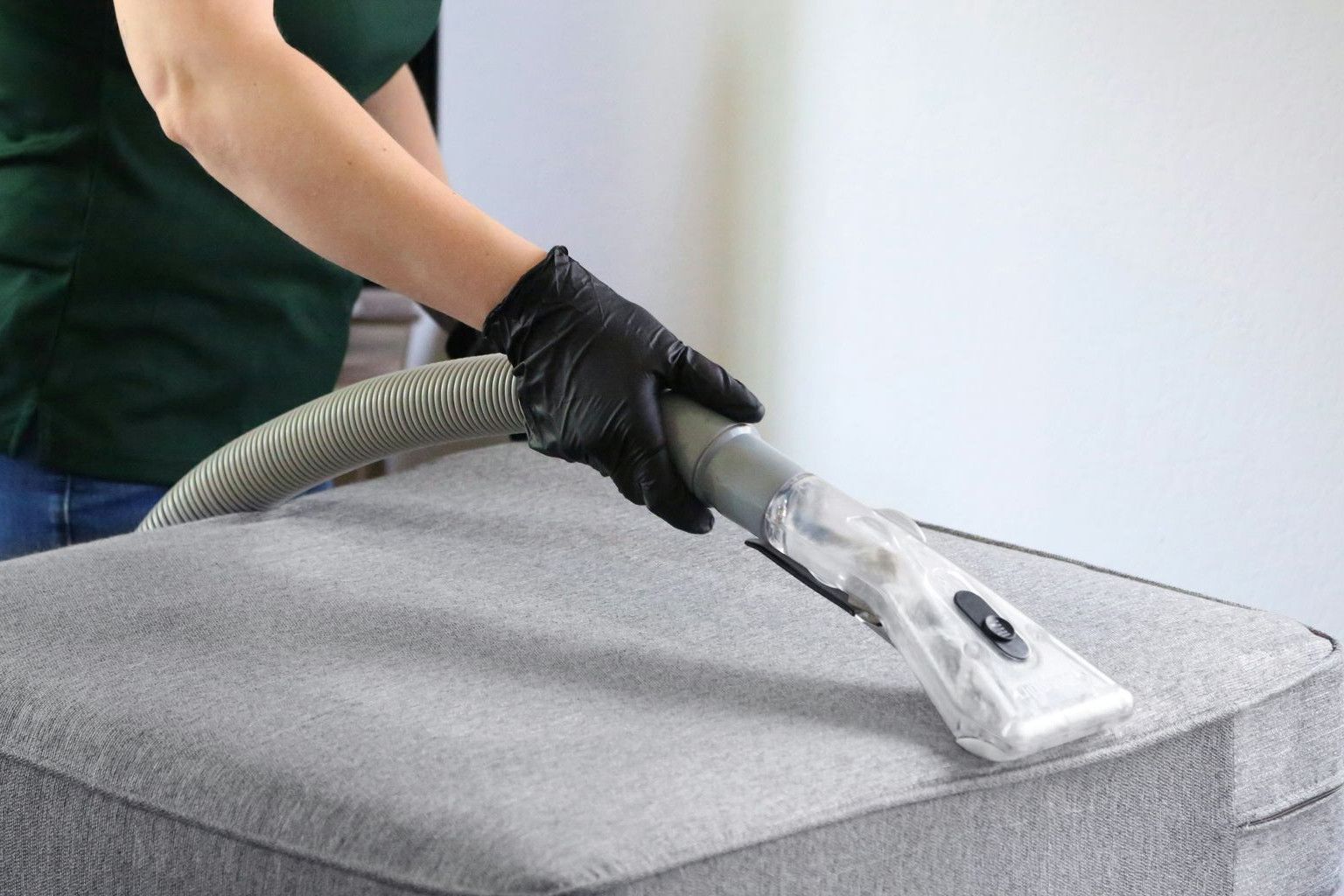 a woman in a green shirt and black gloves vacuuming a gray ottoman